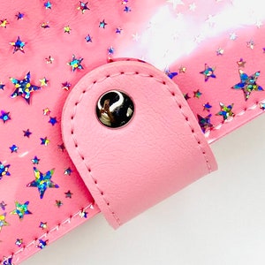 May include: A pink leather binder with a star pattern and a silver metal snap closure.
