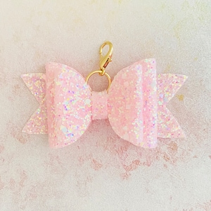 May include: A pink glitter bow with a gold metal clip. The bow is attached to a gold metal ring.