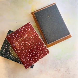May include: Three notebooks with star patterns and a glittery cover. One notebook is burgundy with white stars, another is dark green with silver stars, and the third is black with gold text and the year 2026.