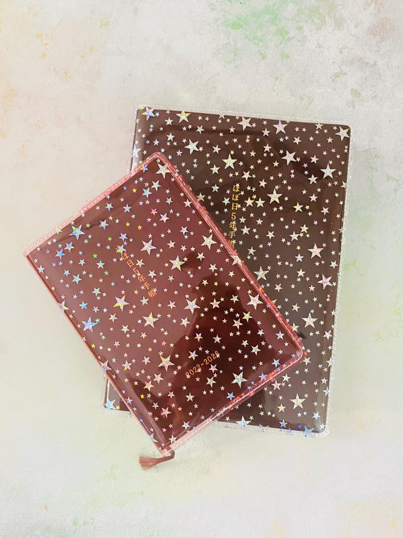 May include: Two brown star-patterned journals with gold foil accents. The smaller journal has the text "2022-2023" printed on the cover. The larger journal has the text "2023年5月始まり" printed on the cover.