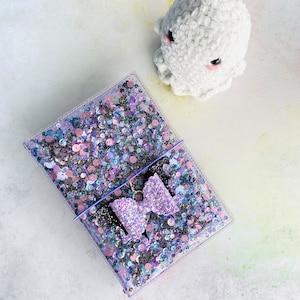 以下が含まれることがあります： 紫のリボンと虹色のスパンコールが付いた透明なプラスチック製のノート。ノートは白い表面に置かれ、背景には白いふわふわの玩具がある。