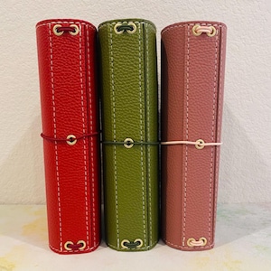 May include: Three vertical leather journals with textured surfaces in red, olive green, and dusty rose. Each journal features white contrast stitching, gold-toned metal eyelets at the top and bottom, and a matching elastic cord closure wrapped around the center.