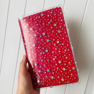 May include: A clear plastic notebook cover with a red background and a pattern of silver glitter stars.