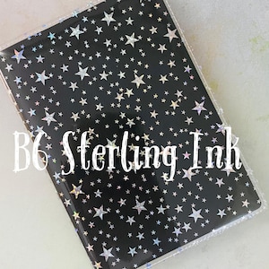 May include: A black notebook cover with a holographic silver star pattern. The cover is clear and has a B6 Sterling Ink logo.