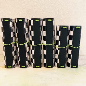 May include: Six journals with a black and white chequered design, black spines, and neon green elastic closures. The journals are displayed in a row, highlighting their distinctive pattern. The covers have a striking checkerboard pattern.