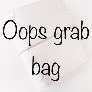 May include: A white grab bag with the words "Oops grab bag" in black lettering. The bag appears to be made of a soft material and is rectangular in shape.