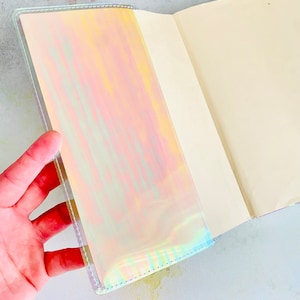 Op de afbeelding: Een holografisch notitieboek met een witte omslag en een open blanco pagina. Het notitieboek heeft een iriserende afwerking die van kleur verandert in het licht.