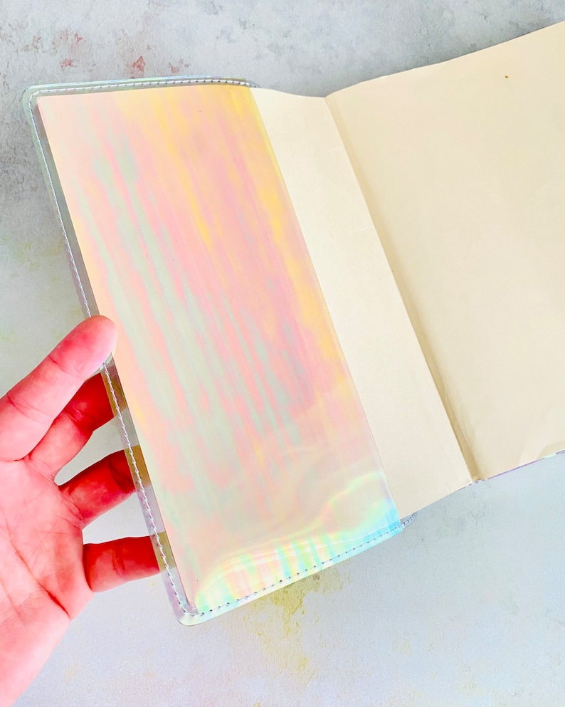 Op de afbeelding: Een holografisch notitieboek met een witte omslag en een open blanco pagina. Het notitieboek heeft een iriserende afwerking die van kleur verandert in het licht.