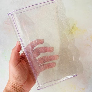 May include: A clear plastic binder with a purple glitter finish and a scalloped edge. The binder has a purple stitching detail around the edges.