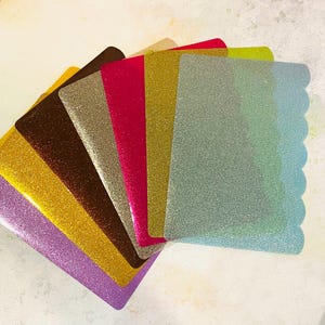 May include: A collection of colorful, glittery card holders fanned out. The colors include purple, gold, brown, silver, red, yellow, and blue-green. The blue-green card holder has a scalloped edge.