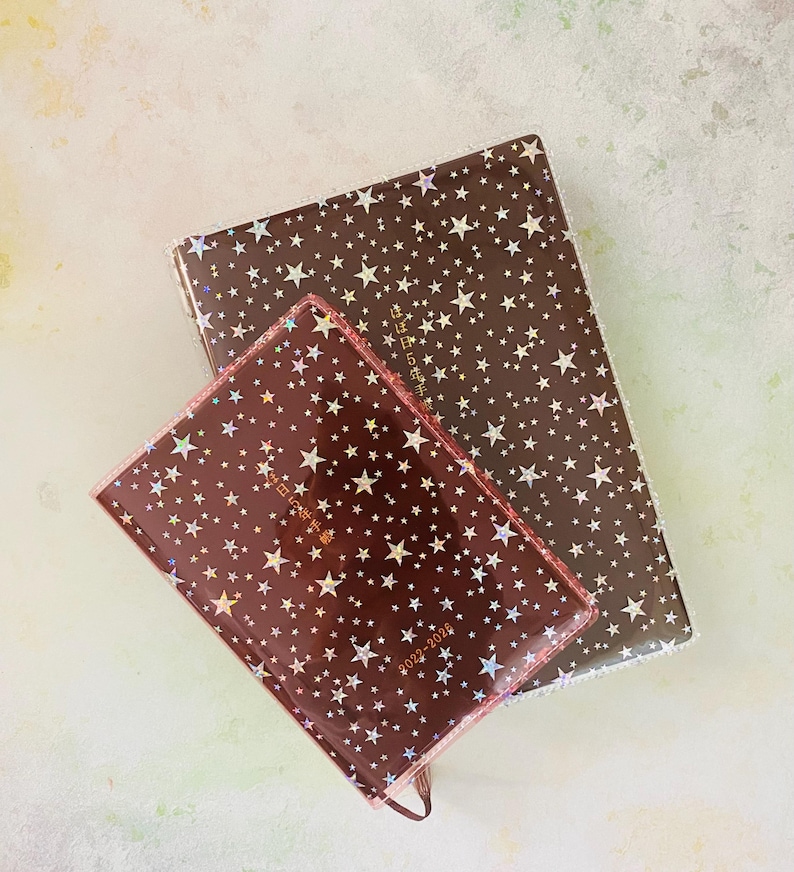 May include: Two brown star-patterned journals with gold foil accents. The smaller journal has the text "2022-2026" and "每日記事手帳" printed on the front. The larger journal has the text "每日記事手帳" printed on the front.