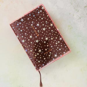 May include: A brown leather-bound journal with a clear plastic cover featuring a holographic star pattern. The cover has the text "2023-2024" and Japanese characters in gold lettering.