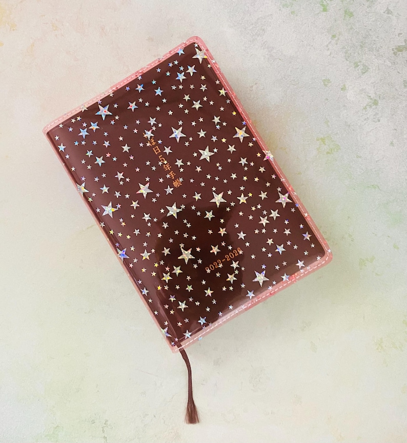 May include: A brown leather-bound journal with a clear plastic cover featuring a holographic star pattern. The cover has the text "2023-2024" and Japanese characters in gold lettering.