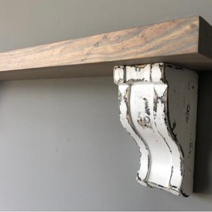 May include: A rustic wooden shelf with a distressed white corbel support. The shelf is stained a light gray color, and the corbel has a worn, vintage appearance. A simple, yet elegant design.