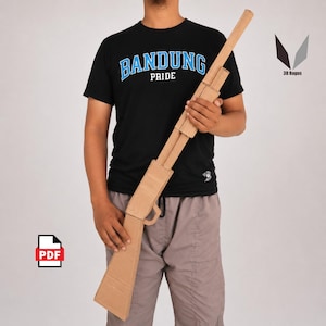 Op de afbeelding: Een kartonnen geweer, vastgehouden door een persoon. Het geweer is gemaakt van golfkarton en heeft een bruine kleur. De persoon draagt een zwart t-shirt met de tekst "BANDUNG PRIDE".