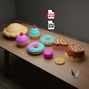 Op de afbeelding: Een verzameling kleurrijke papieren baksels, waaronder een taart, cupcakes, donuts, een macaron, een koekje en een stuk taart. De baksels zijn op een houten tafel gerangschikt. De afbeelding bevat de tekst "PDF" en "SVG".
