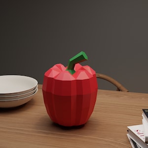 May include: A red and green geometric paper model of a bell pepper on a wooden table with a stack of white plates and a stack of books.