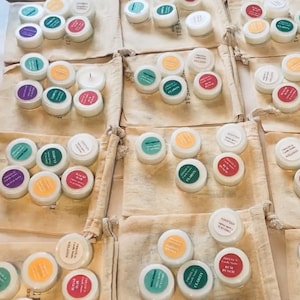 May include: An overhead shot of multiple small, white candle jars with colorful labels, arranged on a white surface and inside beige drawstring bags. The labels feature text like "Clarity" and "Melanin".