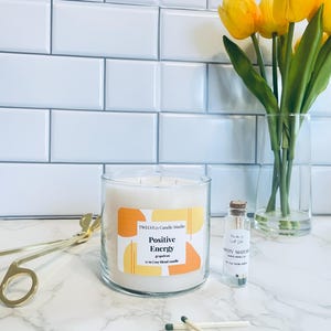 May include: A glass candle jar with a white soy candle inside. The candle jar has a label that says "TWELVE/N Candle Studio" and "Positive Energy" with a grapefruit scent. The candle is 27 oz and is on a white marble surface with a glass jar of matches and a pair of gold tongs.