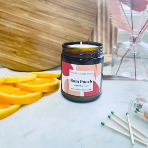 May include: A brown glass candle jar with a white label that reads "TWELVE:21 Candle Studio" and "Rum Punch - Tropical fruit + rum". The candle is on a white surface with orange slices and matches.