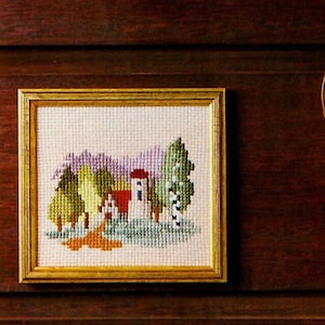 May include: A framed cross-stitch landscape featuring a house with a red roof, surrounded by green, yellow, and purple trees. The artwork is set in a gold-colored frame. The background is a dark wood surface.
