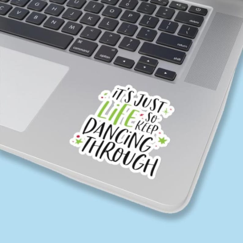Puede incluir: Una pegatina de vinilo troquelada con el texto en negro y verde lima "IT'S JUST LIFE SO KEEP DANCING THROUGH" con peque&ntilde;os detalles de estrellas y puntos rojos y verdes. La pegatina est&aacute; adherida a la superficie plateada de un port&aacute;til.
