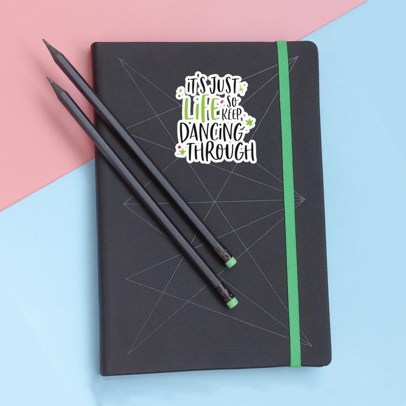 Puede incluir: Un cuaderno negro con una banda el&aacute;stica verde y un patr&oacute;n de estrella geom&eacute;trica en la cubierta, que incluye una pegatina con el texto IT'S JUST LIFE SO KEEP DANCING THROUGH en letras negras y verdes, junto a dos l&aacute;pices gris oscuro con gomas verdes.