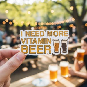 May include: A sticker with the text "I NEED MORE VITAMIN BEER" in a bold, outlined font. The sticker also features two beer glasses. The background is a blurred outdoor scene with people and string lights.