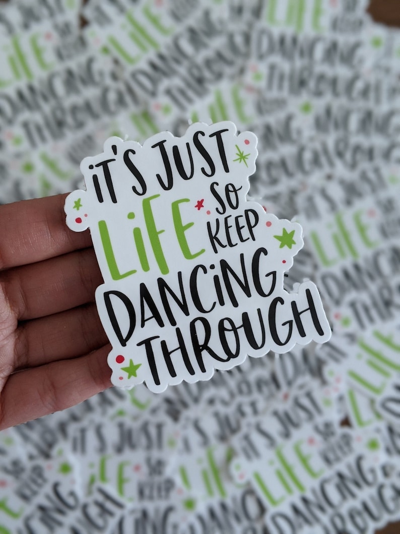 Puede incluir: Una pegatina troquelada con el texto en negro y verde lima IT'S JUST LIFE SO KEEP DANCING THROUGH. El dise&ntilde;o incluye peque&ntilde;as estrellas de color verde lima y puntos rojos dispersos alrededor de las letras sobre un fondo blanco con borde festoneado.
