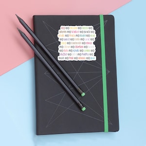 May include: A black notebook with a green elastic band and a white sticker featuring the text: RED AND YELLOW AND GREEN AND BROWN AND SCARLET AND BLACK AND OCHRE AND PEACH AND RUBY AND OLIVE AND VIOLET AND FAWN AND LILAC AND GOLD AND CHOCOLATE AND MAUVE AND CREAM AND CRIMSON AND SILVER AND ROSE AND AZURE AND LEMON AND RUSSET AND GREY AND PURPLE AND WHITE AND PINK AND ORANGE AND BLUE. Two dark grey pencils lie across the notebook.
