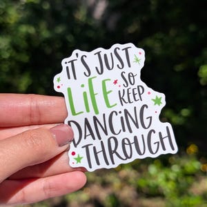 Puede incluir: Una pegatina troquelada con fondo blanco que presenta el texto "IT'S JUST LIFE SO KEEP DANCING THROUGH" en una mezcla de letras negras y verdes. El dise&ntilde;o est&aacute; decorado con peque&ntilde;as estrellas verdes y puntos rojos dispersos.