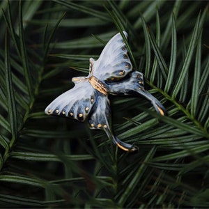 Può includere: Primo piano di un oggetto decorativo a forma di farfalla. La farfalla ha ali bianche e blu con dettagli dorati, su uno sfondo di fogliame verde. Il design intricato dell'oggetto è evidenziato.