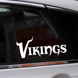 Pode incluir: Um adesivo de vinil branco com a palavra "Vikings" em uma fonte estilizada, aplicado na janela traseira de um carro.