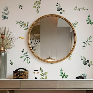 Op de afbeelding: Een ronde spiegel met een gouden lijst op een witte muur die is versierd met groene bladeren. Een houten kaptafel met twee lades staat op de voorgrond.