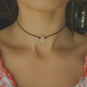 Może przedstawiać: Czarny naszyjnik typu choker z przezroczystym kryształowym wisiorkiem. Kryształ wisi na cienkim czarnym sznurku. Osoba ma na sobie top w różowo-biały wzór.