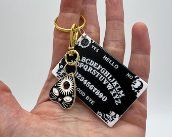 May include: A black and white Ouija board keychain with a gold-colored metal ring and a black and white cat-shaped charm with a sun and moon design. The keychain features the letters of the alphabet, numbers 1-0, and the words "YES", "NO", "HELLO", and "GOOD BYE".