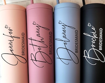 May include: Four tall, cylindrical tumblers with matching straws in matte peach, dusty rose, light blue, and black. Each tumbler features a custom name in black script, a small heart icon above the name, and the word BRIDESMAID in block letters.