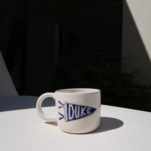 May include: White ceramic mug with a curved handle. The mug features a blue pennant design with the word "DUKE" printed on it. The mug is sitting on a white surface.
