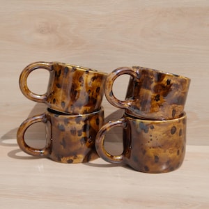 May include: Four ceramic mugs arranged in a stack, each featuring a glossy, mottled brown and black tortoiseshell pattern. The mugs have rounded bodies and curved handles, with a smooth, reflective finish across their entire exterior surface.