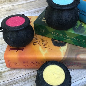 Peut inclure: Trois bombes de bain en forme de chaudron noir avec des sels de bain de différentes couleurs à l'intérieur. Les bombes de bain sont posées sur des livres Harry Potter.