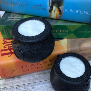 May include: Two black cauldron-shaped candle holders with white wax inside, sitting on top of a stack of Harry Potter books. The top book is "Harry Potter and the Goblet of Fire" by J.K. Rowling.