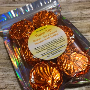 May include: A bag of orange and lemon scented shower steamers with menthol. The bag is labeled "Wake Up! Orange - Lemon Shower Steamers With Menthol".