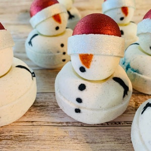 May include: A collection of snowman-shaped bath bombs. Each white bath bomb has a red glittery hat, a white brim, a carrot nose, and black button eyes and mouth. The bath bombs are arranged on a light wood surface.