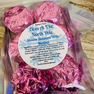 May include: A clear plastic bag filled with several pink foil-wrapped shower steamers. The label reads "Elves At The North Pole Shower Steamers With Menthol" and lists ingredients and usage instructions. The Etsy shop name "JustBubblesAndBrews" is also visible.