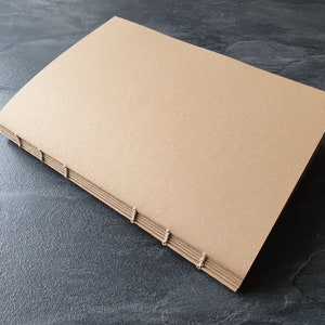 Könnte beinhalten: Ein handgenähtes Notizbuch in Beige mit einer strukturierten Oberfläche. Die Seiten sind mit sichtbarem Faden gebunden, was eine rustikale und handwerkliche Ästhetik erzeugt. Das Notizbuch liegt offen auf einer dunkelgrauen Oberfläche.