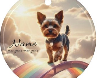 May include: Round ceramic ornament featuring a Yorkshire Terrier dog walking on a rainbow bridge against a cloudy sky background. The ornament has the text "Name" and "mo/year-mo/year". A gold string is attached for hanging.