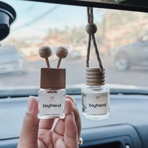 以下が含まれることがあります： 木製の蓋と透明なガラス瓶が付いた車の芳香剤が2つ。両方の瓶にはピンクのハートと「boyfriend」という文字がラベルに書かれています。1つの芳香剤はルームミラーに吊り下げられており、もう1つは手で持たれています。