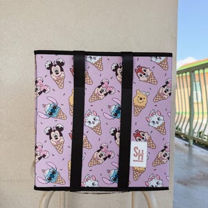 May include: A rectangular tote bag with a light purple background featuring cartoon characters on ice cream cones. The bag has black handles and trim. A small white label with "SH" is visible.