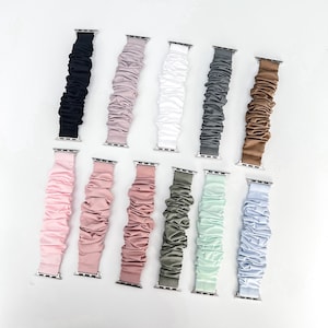 May include: Eleven scrunchie-style watch bands arranged in two rows. The colors include black, mauve, white, charcoal, tan, light pink, dusty rose, salmon, olive green, mint green, and light blue. Each band features metal connectors.