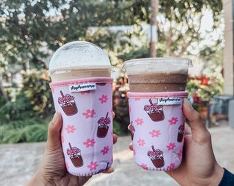May include: Two pink and white insulated beverage sleeves with a floral pattern and a cartoon image of a glass of iced coffee. The text "Sip & Savor" is printed on the sleeves.